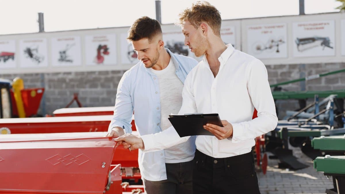 Two men discussing agricultural equipment purchase at a dealership, one holding a clipboard outside.