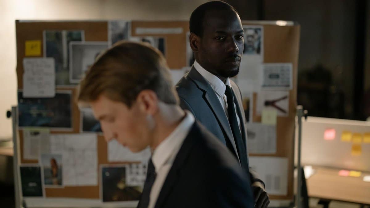 Two detectives examining a corkboard filled with evidence in a dimly lit office.