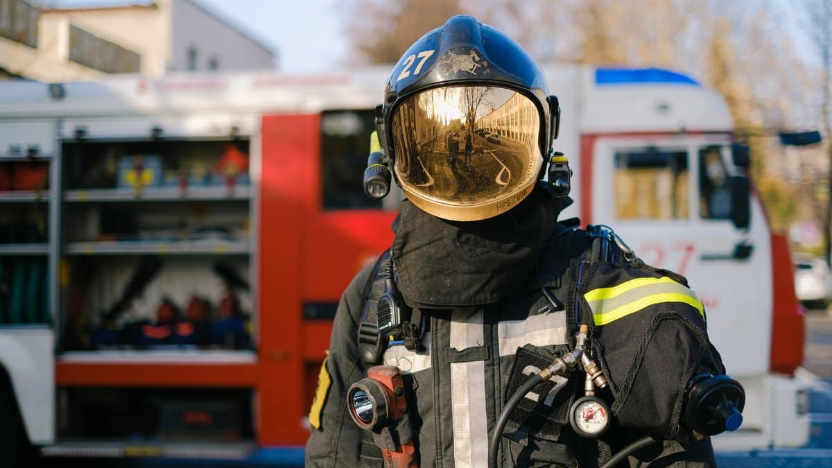 Firefighter in full protective gear with a reflective helmet stands in front of a fire truck on a sunny day.