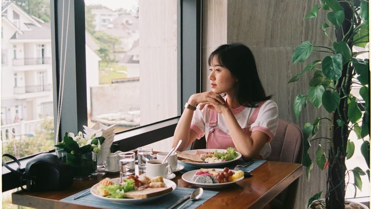 A woman enjoys a meal by a window, thoughtfully gazing outside in a bright restaurant setting.