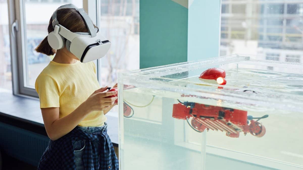 Side view of little girl wearing VR goggles and exploring new robot with controllers in light room