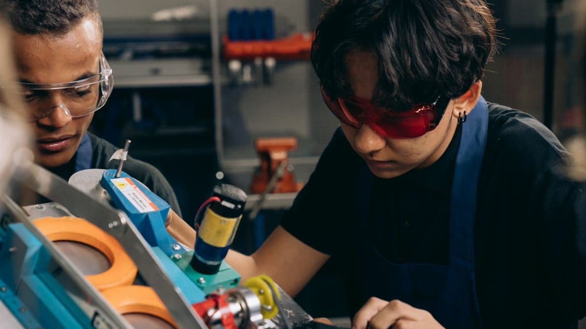 Two young engineers concentrating on precision work in a technical workshop, wearing safety goggles.