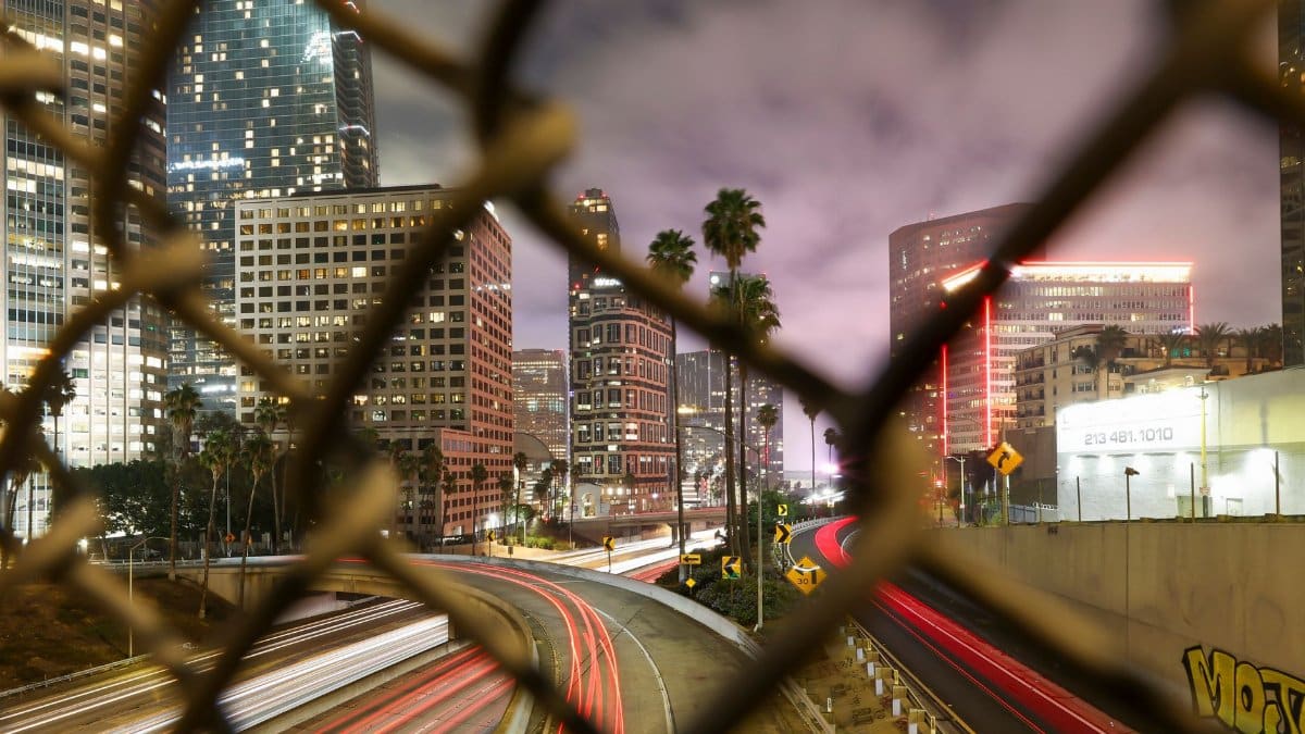 Long exposure night shot capturing the dynamic Los Angeles cityscape through a chain-link fence.