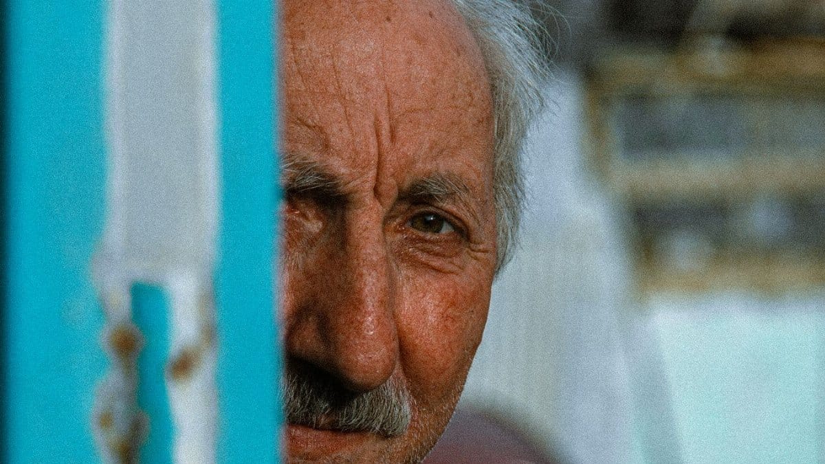 Close-up portrait of an elderly man with a mustache peering from behind a wall.