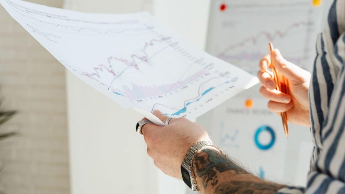 Close-up of a tattooed man's hands holding financial charts, with focus on data analysis and stock market trends.