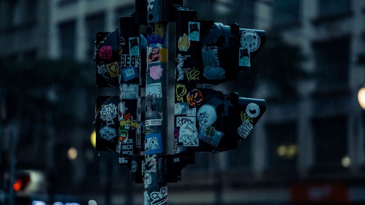 A dimly lit urban traffic light adorned with various street art stickers during a rainy night.