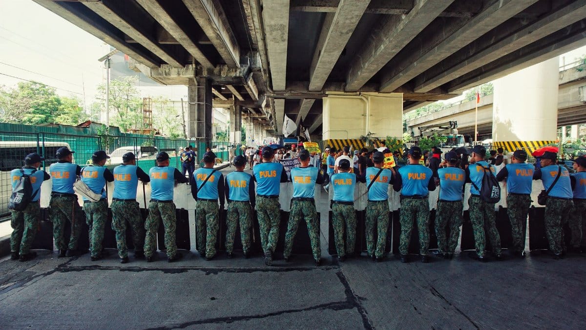 A line of police officers in blue uniforms standing under a bridge during a daylight operation.