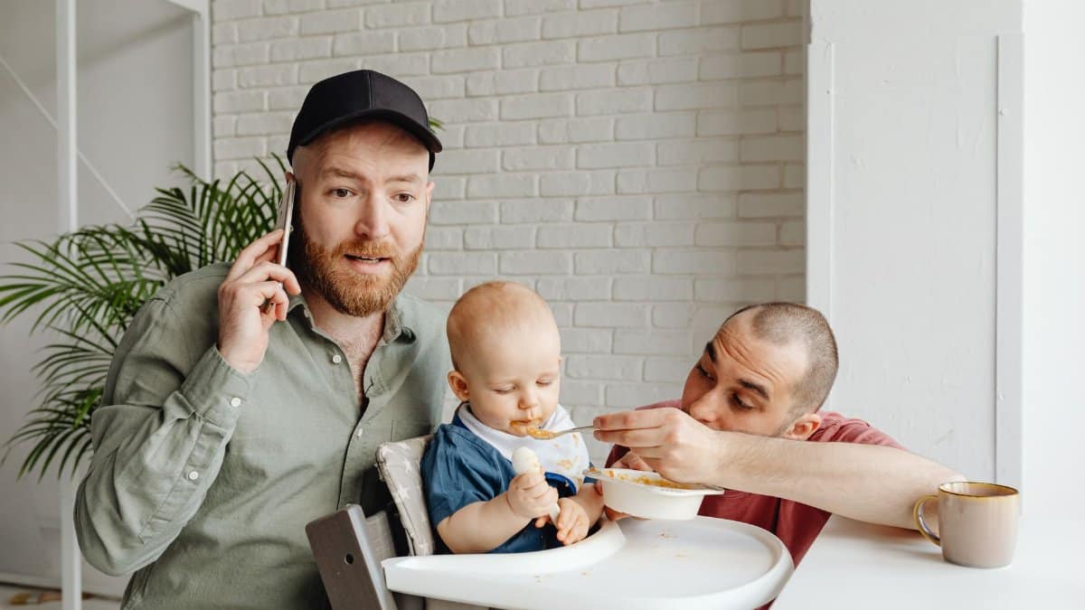 Two dads feeding their baby in a high chair, celebrating family and love indoors.
