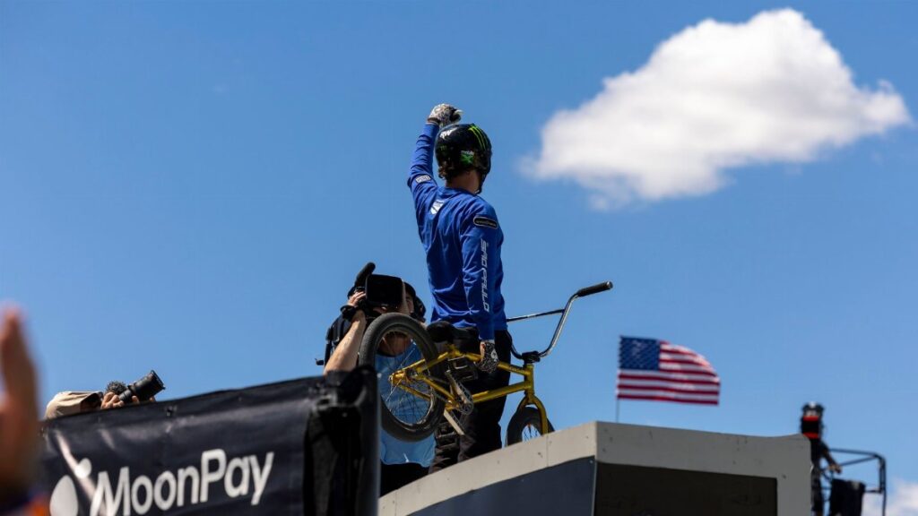 BMX rider celebrates atop ramp with American flag and clear sky, highlighting extreme sports excitement.