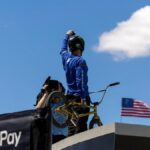 BMX rider celebrates atop ramp with American flag and clear sky, highlighting extreme sports excitement.
