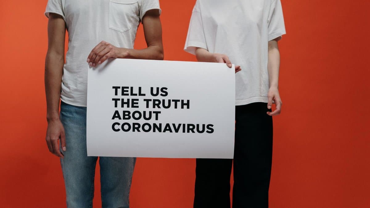 Two individuals holding sign demanding truth about coronavirus against orange background.
