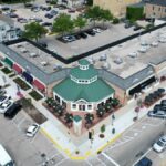 Bird's-eye view of a bustling suburban shopping center with parked cars and tree-lined streets.