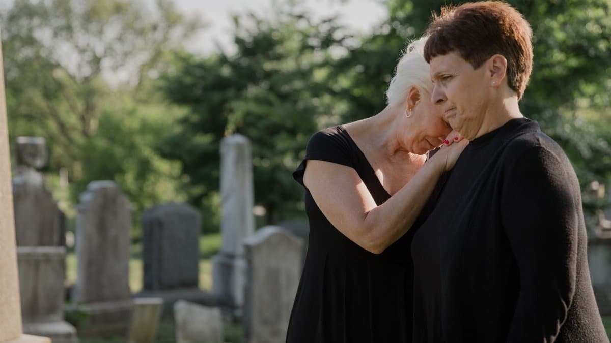 Two women share a comforting moment in a serene cemetery setting.