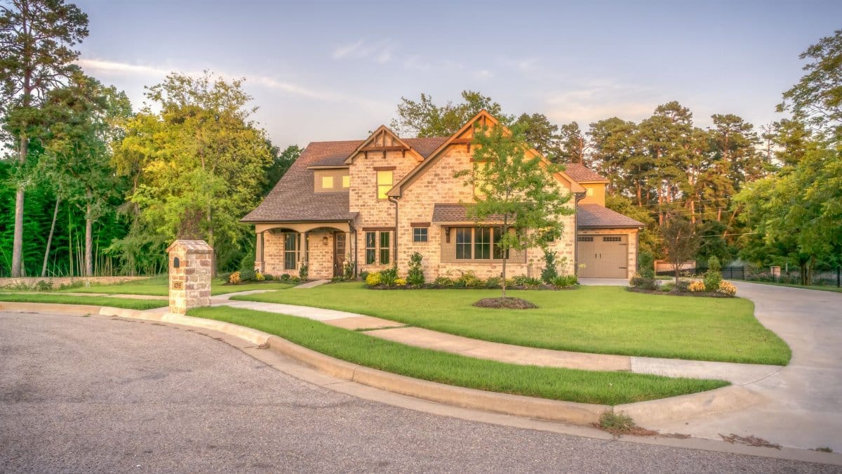 Elegant stone family home with manicured lawn, trees, and driveway on a sunny day.