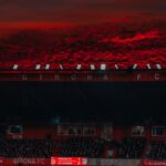 A stadium filled with fans under a dramatic red sunset sky during a Girona FC match.