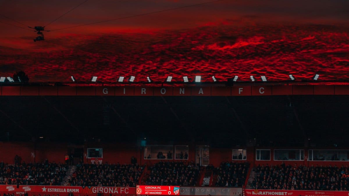 A stadium filled with fans under a dramatic red sunset sky during a Girona FC match.