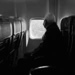 A man wearing a mask gazes out an airplane window during a daytime flight.
