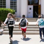 Group of students with backpacks running into school, captured from behind, showing excitement.