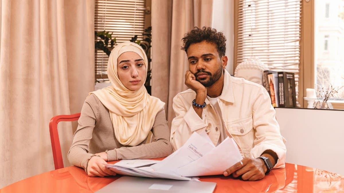 A couple in casual attire sits indoors at a table reviewing financial documents with a laptop.