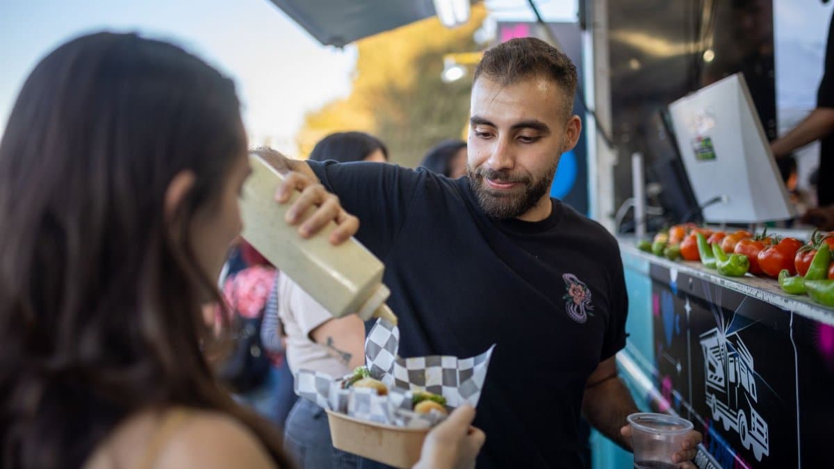 Vendor adds sauce to a customer's dish at an outdoor food truck, featuring fresh peppers and a casual atmosphere.