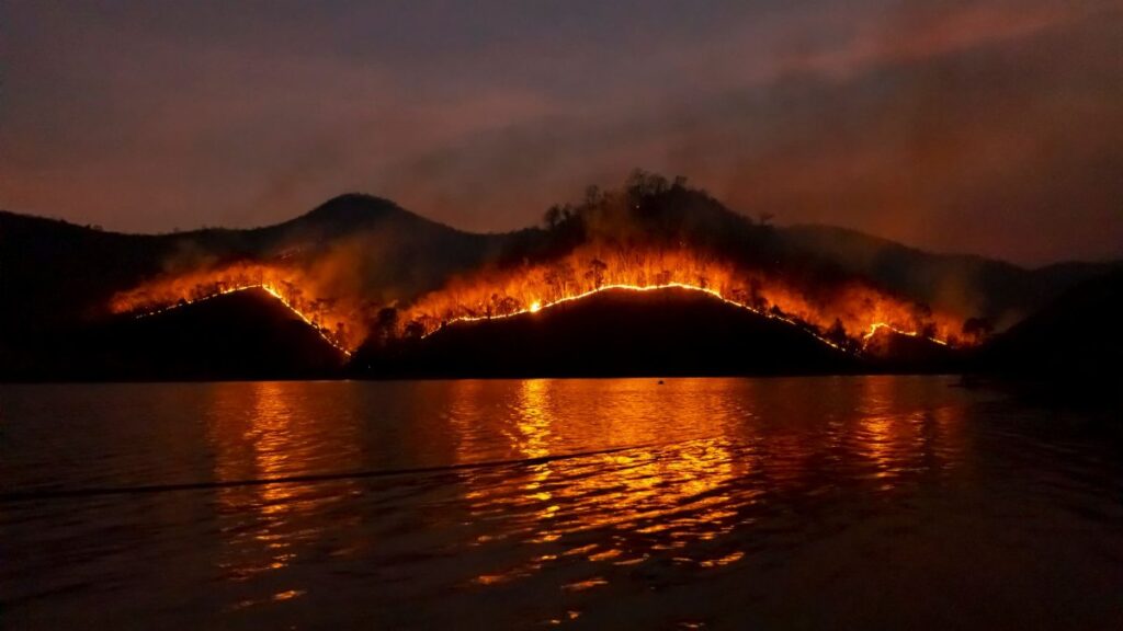 Dramatic wildfire in Thailand with blazing mountains reflecting in the dark waters.