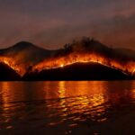 Dramatic wildfire in Thailand with blazing mountains reflecting in the dark waters.