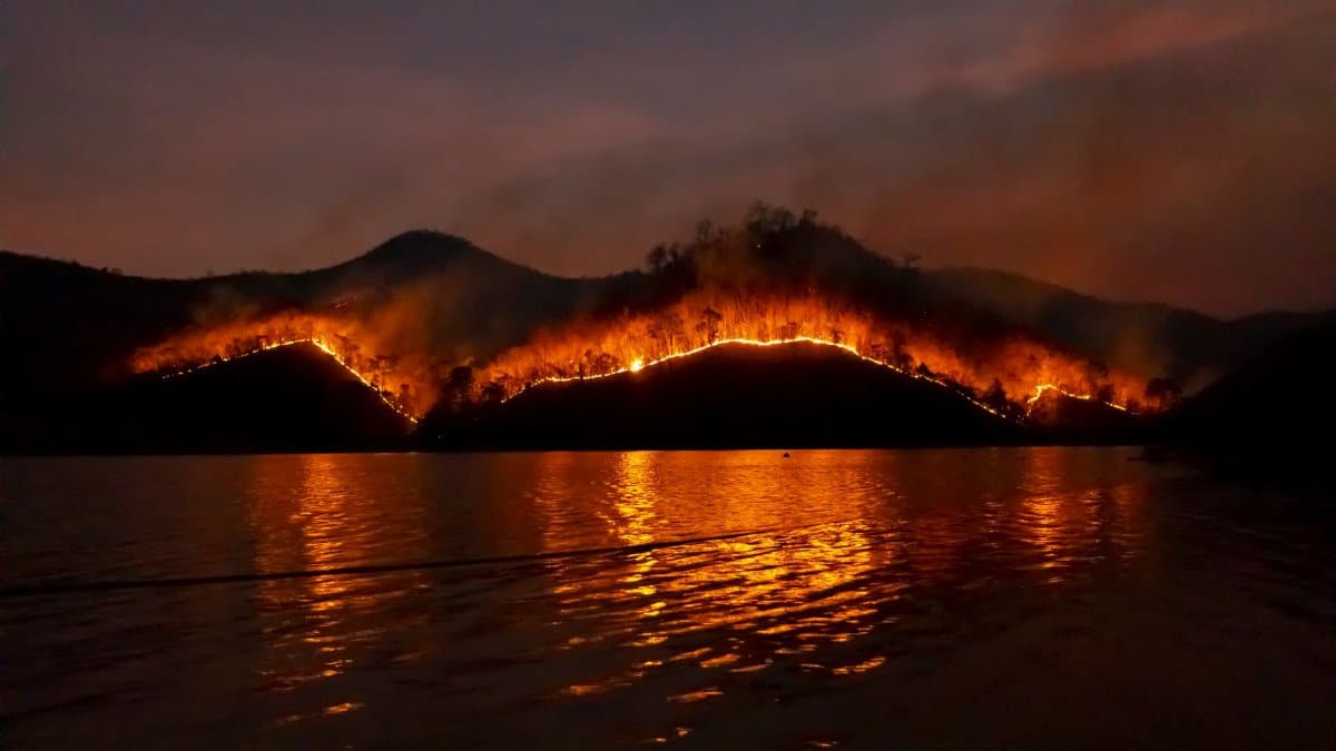 Dramatic wildfire in Thailand with blazing mountains reflecting in the dark waters.