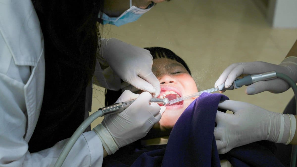 A young child receiving dental treatment from a masked dentist using dental tools indoors.