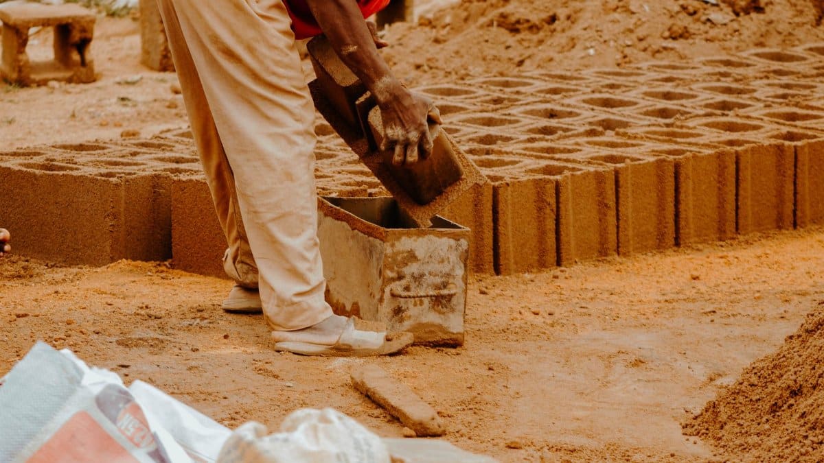 A worker shaping bricks with a concrete mold at an outdoor construction site.