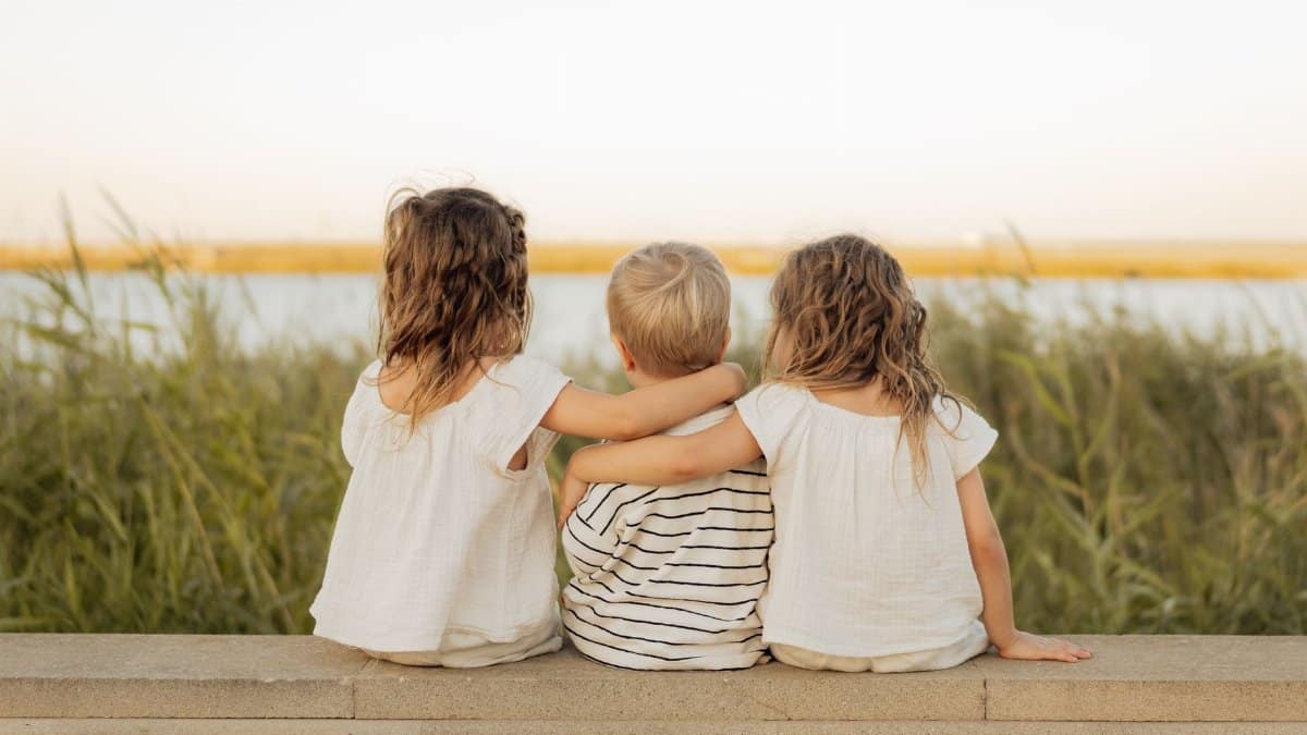 Three children sit on a stone ledge, gazing at a serene lakeside scene, embodying friendship and tranquility.