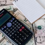From above electronic calculator and notepad placed over United States dollar bills together with metallic pen for budget planning and calculation