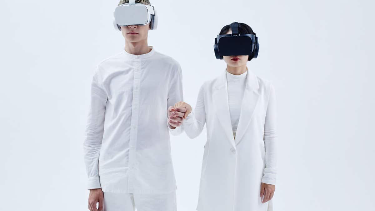 Two people in white attire wearing virtual reality headsets and holding hands, embracing technology.