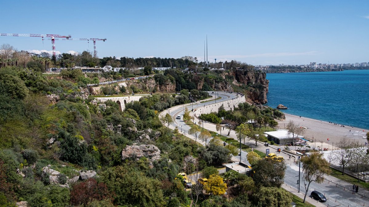 A scenic coastal road overlooking the ocean with lush greenery and a sandy beach on a sunny day.