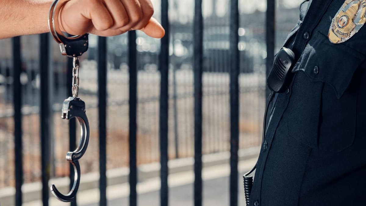 Close-up of a police officer handcuffing an individual against a metal fence outdoors.