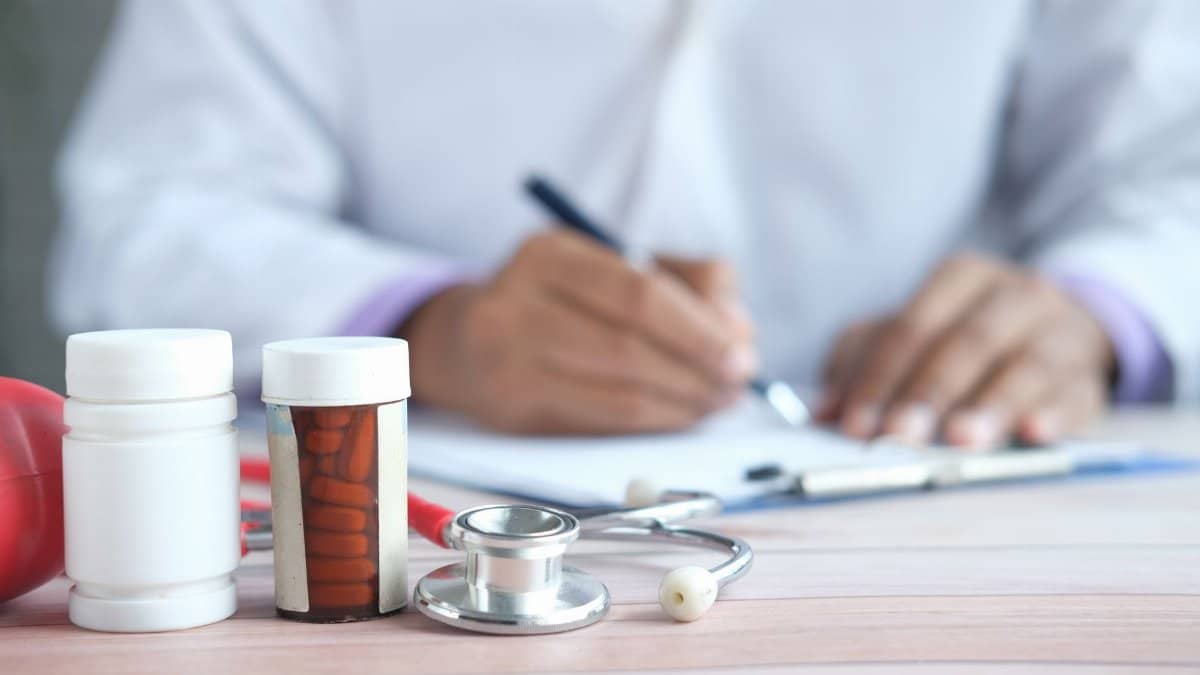Close-up of doctor writing prescription with medicines and stethoscope on desk.