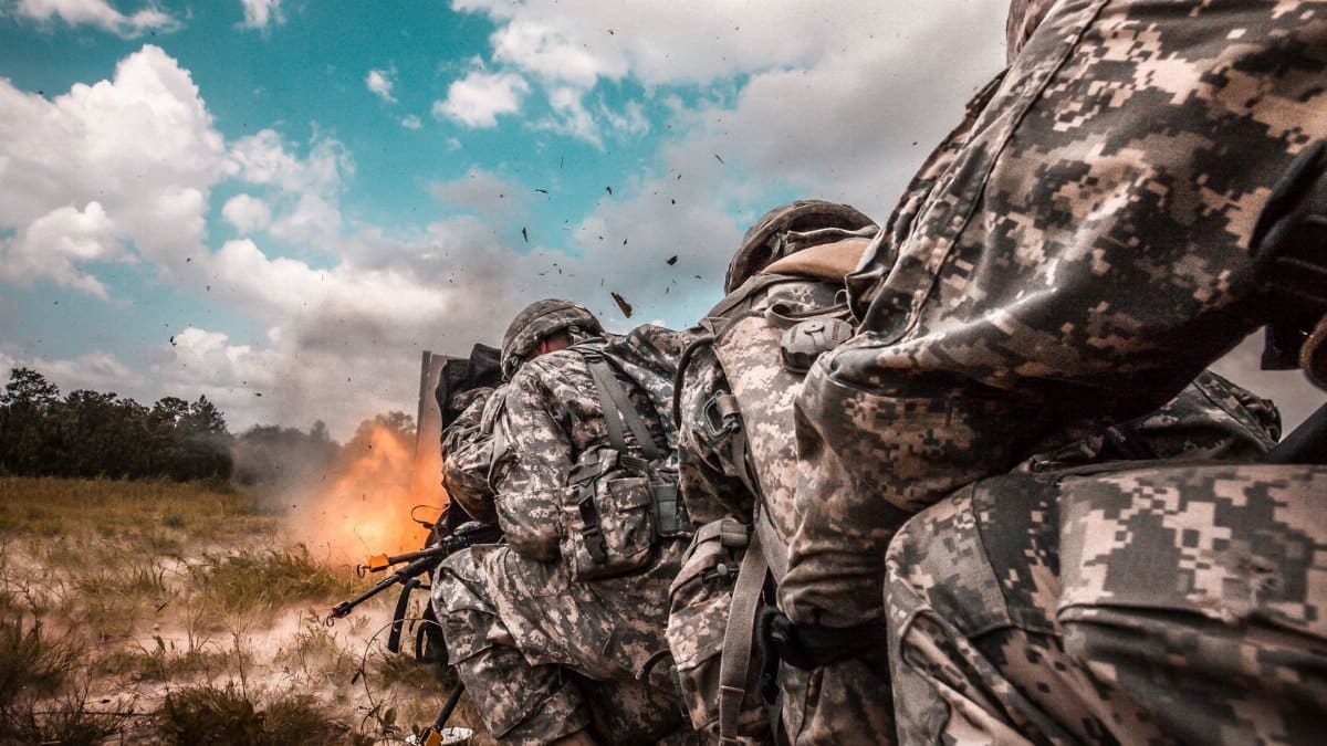 Military soldiers in action during training as explosion occurs in the background.