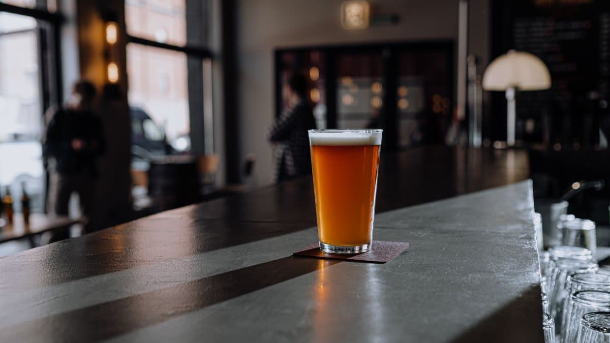 A glass of craft beer with foam sits on a bar counter in a dimly lit modern bar.