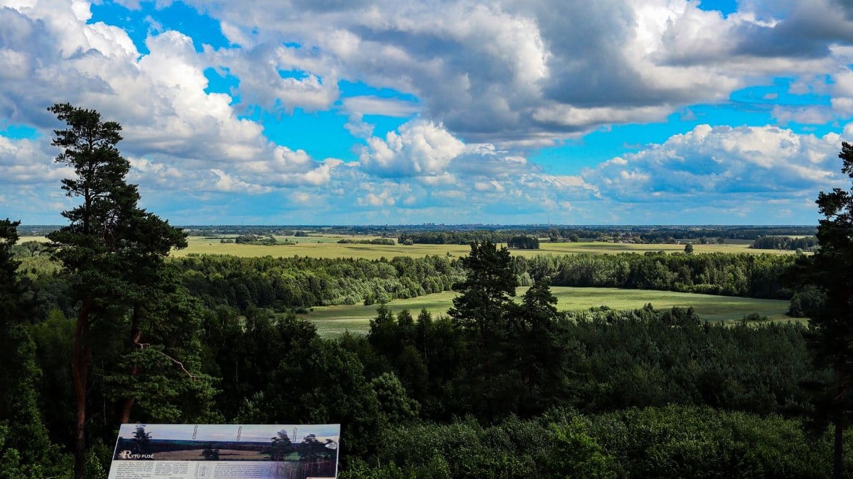 Panoramic landscape of lush green fields and forests beneath a vibrant sky with scattered clouds.