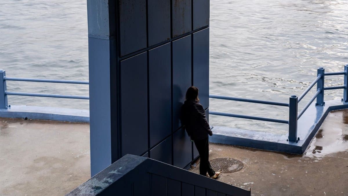 A lone figure stands by a waterfront wall, reflecting at dusk.