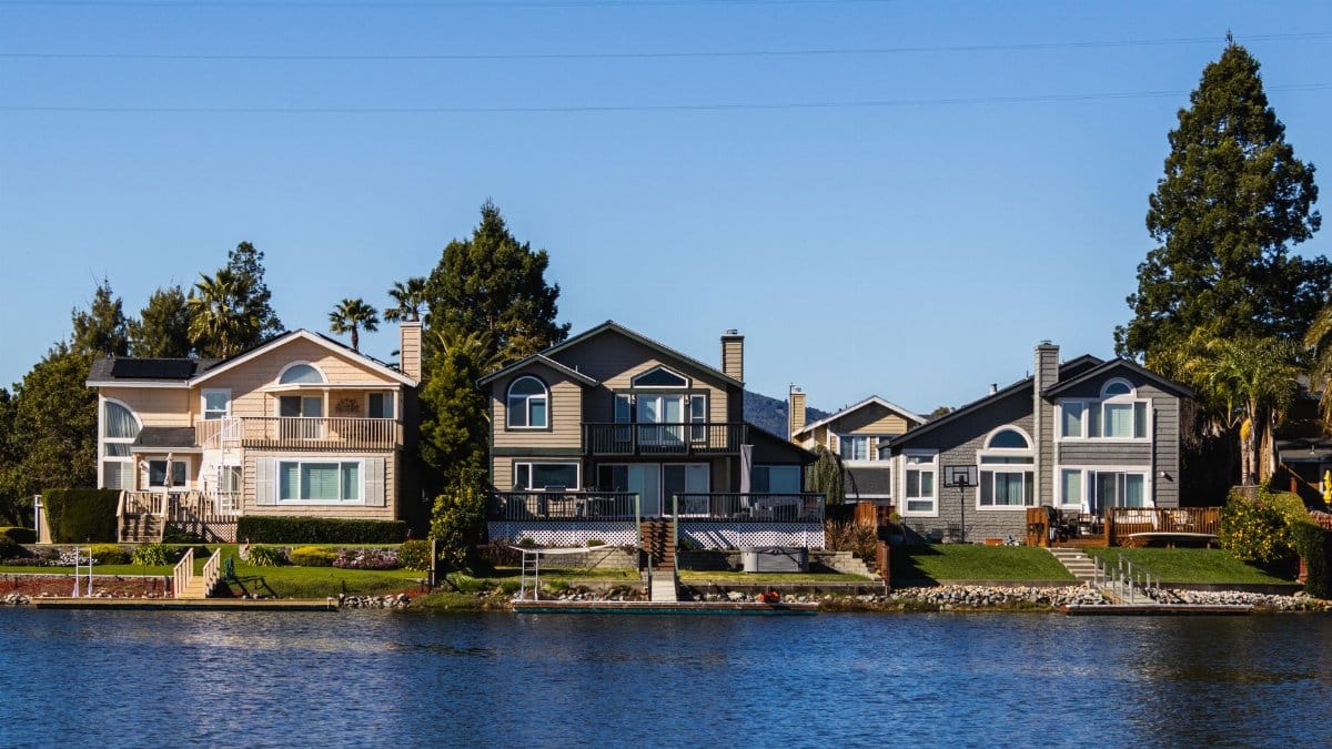 Charming lakeside houses reflect on calm waters under a clear blue sky.