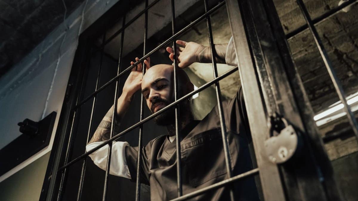 A bald prisoner with tattoos stands in a prison cell, gripping the bars.