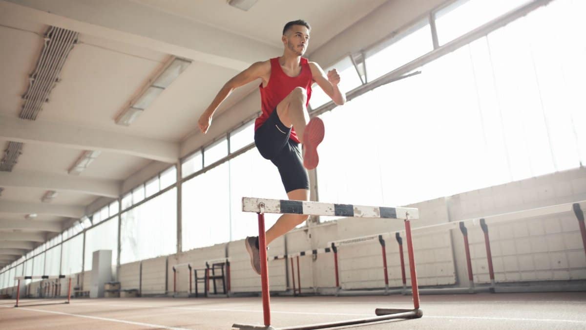 Powerful male athlete jumping over hurdle on running track while training at sports hall