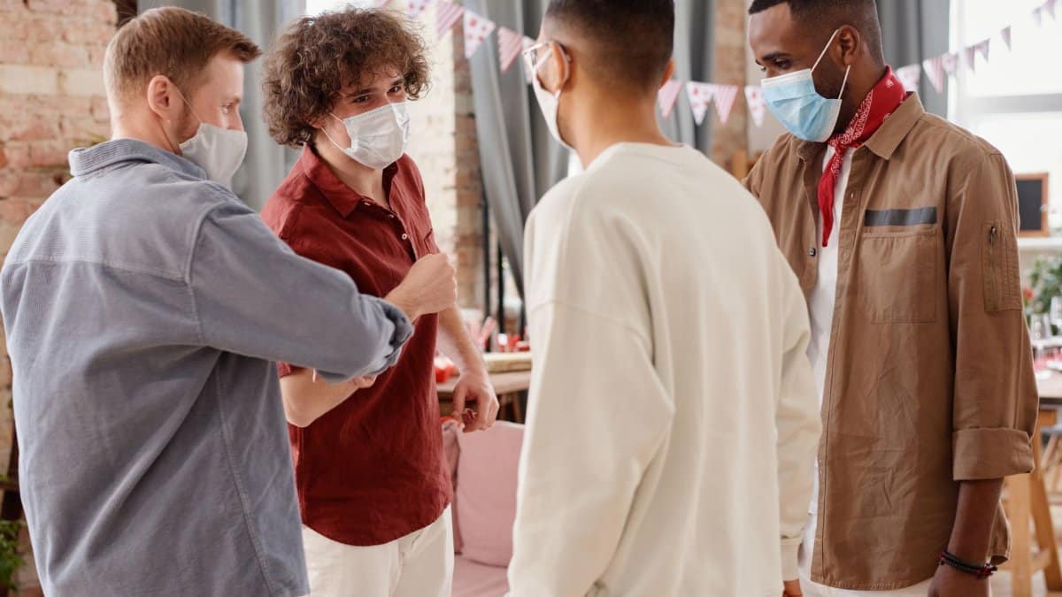 Four diverse men wearing masks socializing inside, emphasizing safe gatherings.