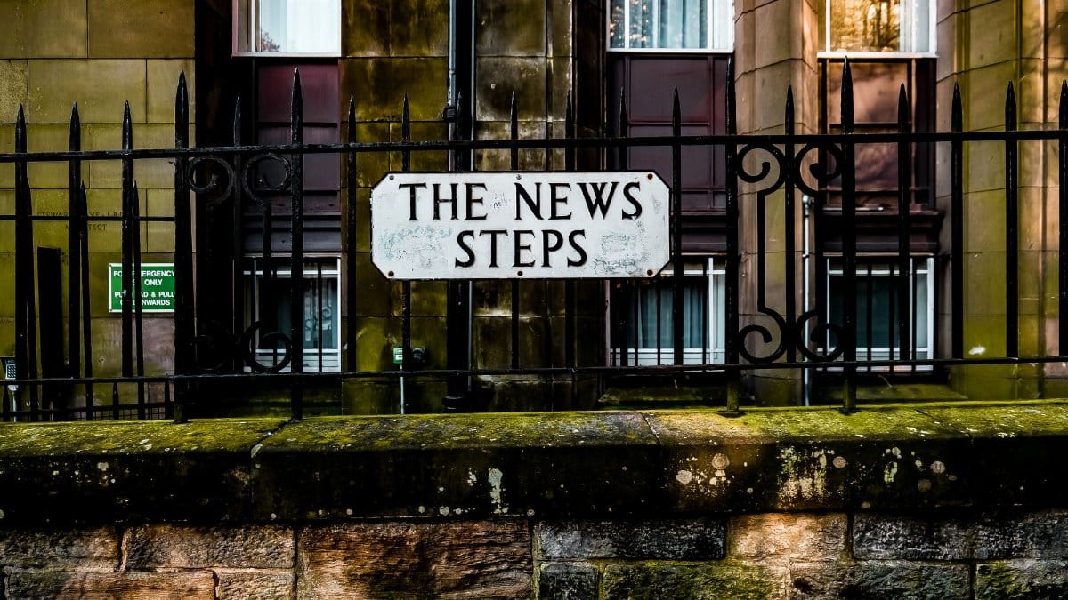 The News Steps sign on a historic iron fence with stone building backdrop.