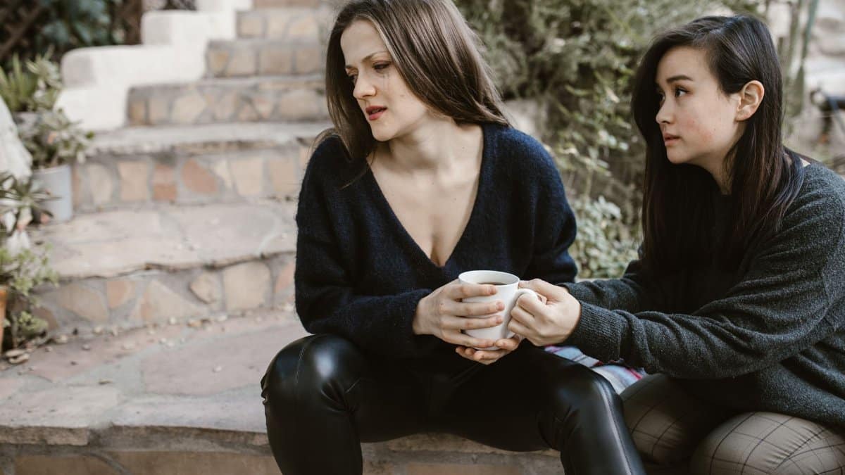 Two women share emotional support on outdoor steps, one consoling the other with a cup.