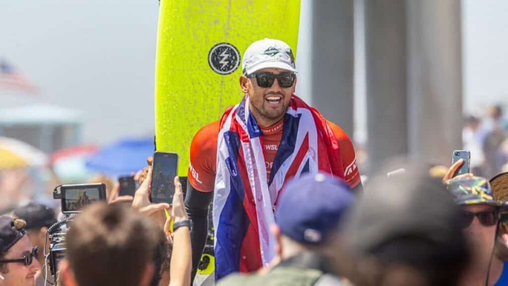 Surfing champion celebrates victory wrapped in flag, surrounded by fans at sunny beach event.
