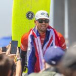 Surfing champion celebrates victory wrapped in flag, surrounded by fans at sunny beach event.