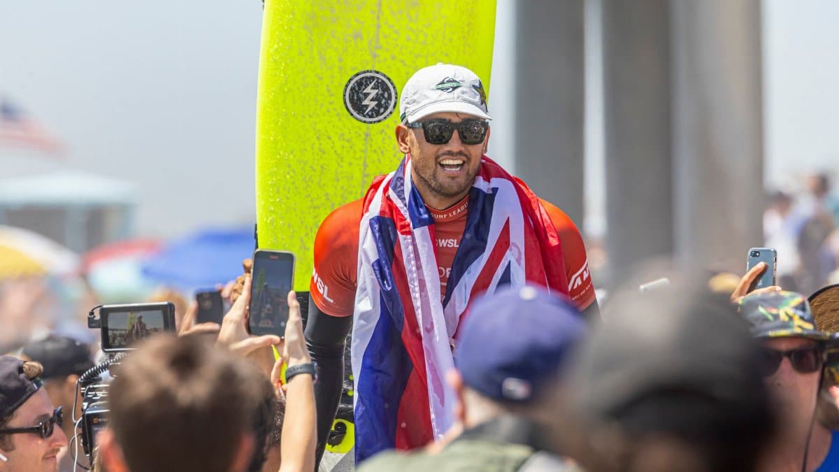 Surfing champion celebrates victory wrapped in flag, surrounded by fans at sunny beach event.