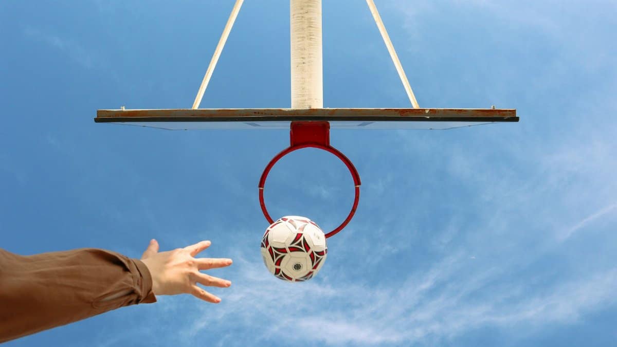 Unique angle capturing a soccer ball nearing a basketball hoop against a bright blue sky.