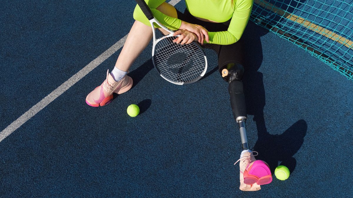 Woman with prosthetic leg resting on tennis court with racket and balls, embracing sport inclusivity.